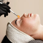 Close-up of a cosmetologist applying a cosmetic peeling mask to a client’s face. The scene emphasizes the professional approach to skin care and the relaxing spa experience.